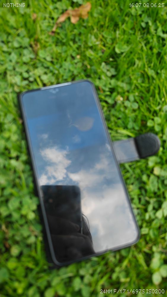 A Samsung Galaxy S21 lies face-up on a clover-covered lawn, reflecting a partly cloudy sky in its dark screen. The phone’s black flip case is open, and the photographer’s faint silhouette is visible in the lower portion of the screen reflection.