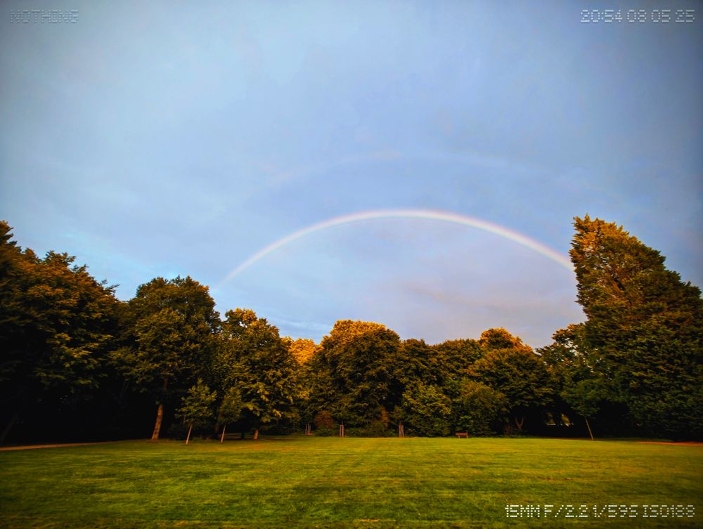 A quiet park beneath a cloudy sky, where a full rainbow arcs gently above the treetops. The grass is lush, the trees hold stillness, and a second, fainter arc echoes above the first. A lone bench rests beneath the signal — untouched, as if waiting for someone aligned enough to sit without breaking it.



