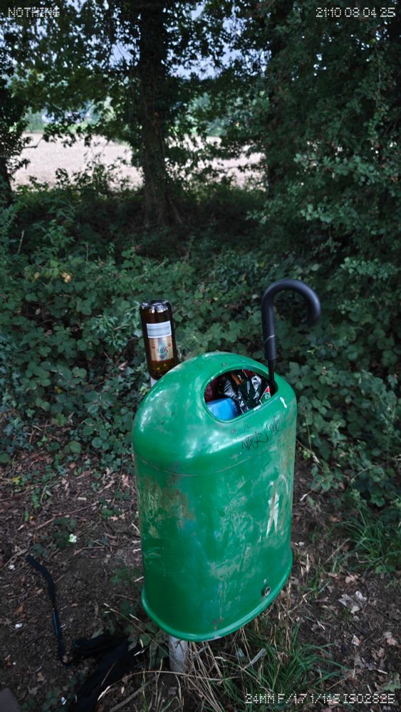 A green forest-side trash bin, overflowing with beer bottles, snack wrappers, and a black umbrella awkwardly stuffed inside. The bin is tagged with graffiti and stands among dense undergrowth. Shot on a Nothing Phone 3 at 24mm f/1.7, the timestamp reads 21:10 08 04 25. The chaotic contents mirror a brief, aimless consumption driven culture. 