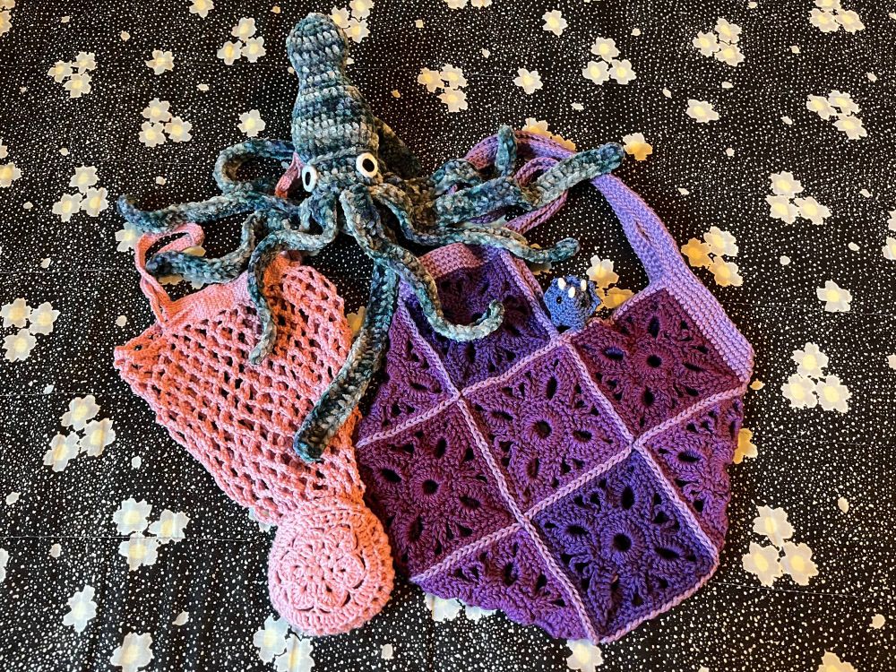 Four crocheted pieces laid out on top of each other: A multicolored chenille squid, a pink mesh tote bag with an attached pouch it fits into, a multi-shade purple granny square tote bag, and a tiny blue triceratops