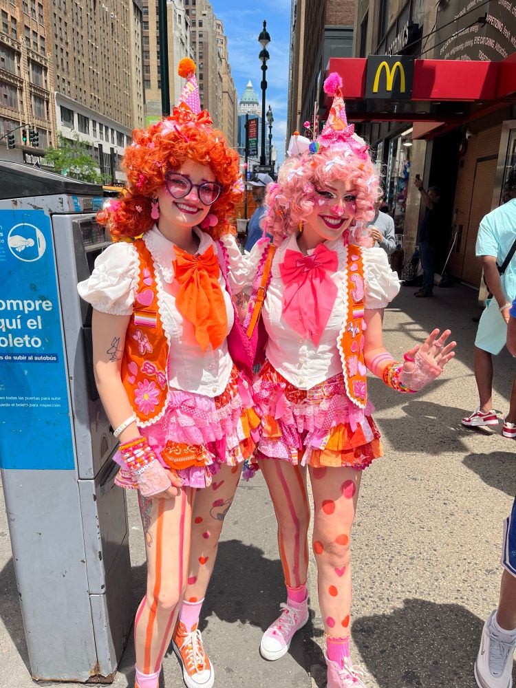 Two nice lesbians smiling big and dressed in colorful matching jester/clown outfits in the colors of the lesbian pride flag. One is wearing an orange wig, the other is wearing a light pink wig