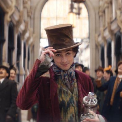 Timmy Chalamet in the upcoming Wonka movie, wearing a brown top hat, a dark red overcoat, and a multicolored vest and scarf