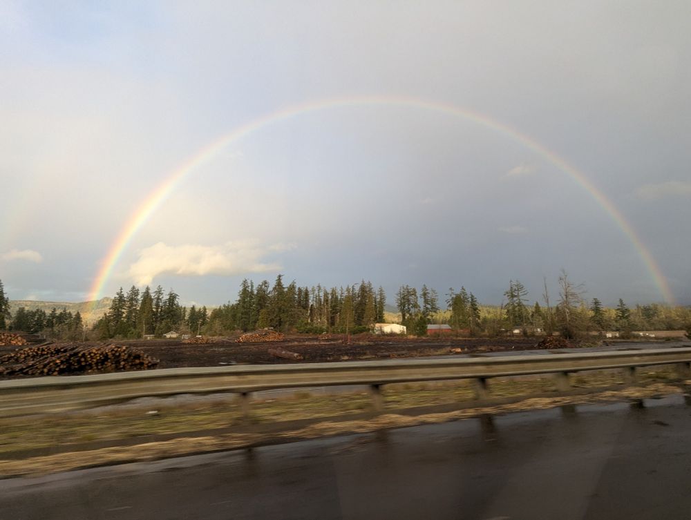 A rainbow stretches across the sky with trees and houses on the horizon.