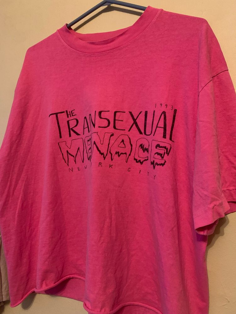 a neon pink cropped t shirt photographed hung up on a wall with the words THE TRANSEXUAL MENACE New York City written off kilter in sharpie on it