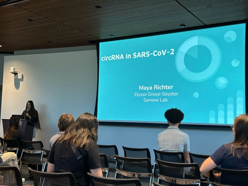 A student stands at a podium on the left of the image as she presents a blue title slide on a large screen, on the right of the image. Audience members sit in the foreground.