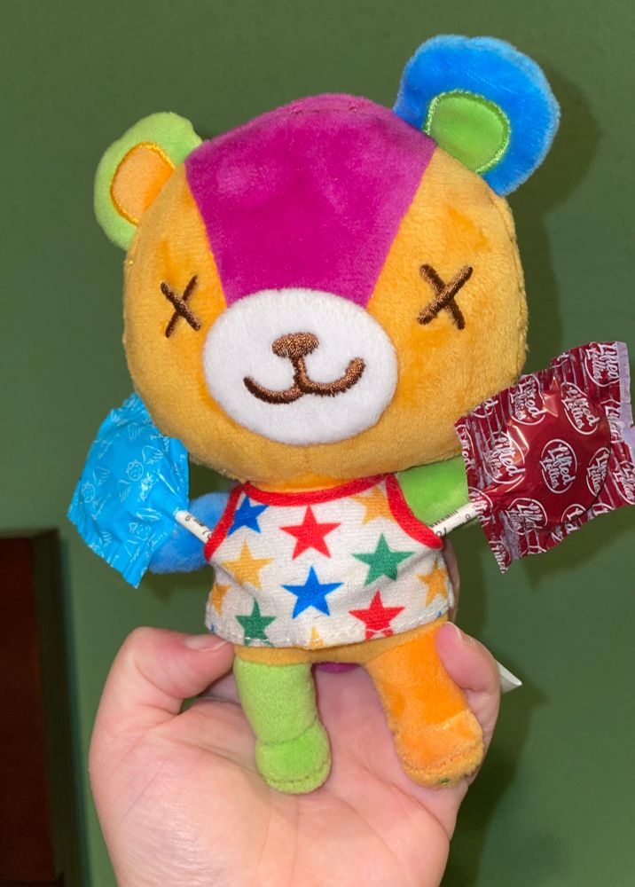 a multicolored bear plushie is wearing a white top with multicolored stars and is holding two garden lollies 