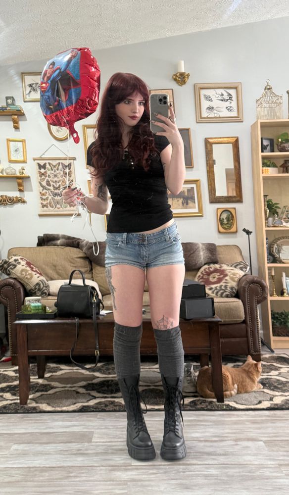 mirror selfie in a black henley top, micro denim shorts, grey knee high socks, and black mid calf combat boots holding up a superman ballon