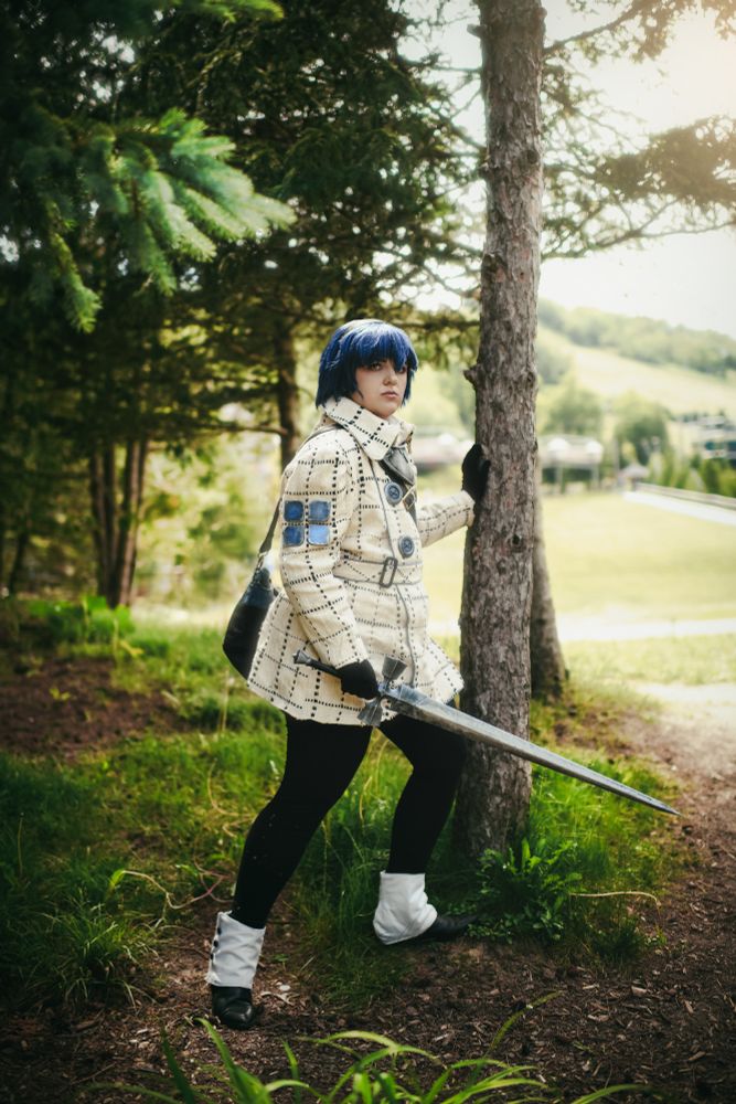 a cosplayer dressed as blue-haired will/the protagonist from metaphor re:fantazio in a white plaid coat with blue accents, leggings, and boots. they're looking out over a hill holding a silver-blue sword.