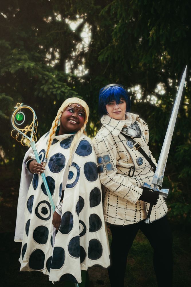 two cosplayers dressed as Eupha, a girl in a black and white dress with blonde hair and a third eye, and Will, in a white double breasted coat with blue buttons and a sword, from Metaphor: re fantazio. They're standing back to back, and Eupha is holding a silver staff and smiling, while Will is holding a silver sword and smirking back at her.