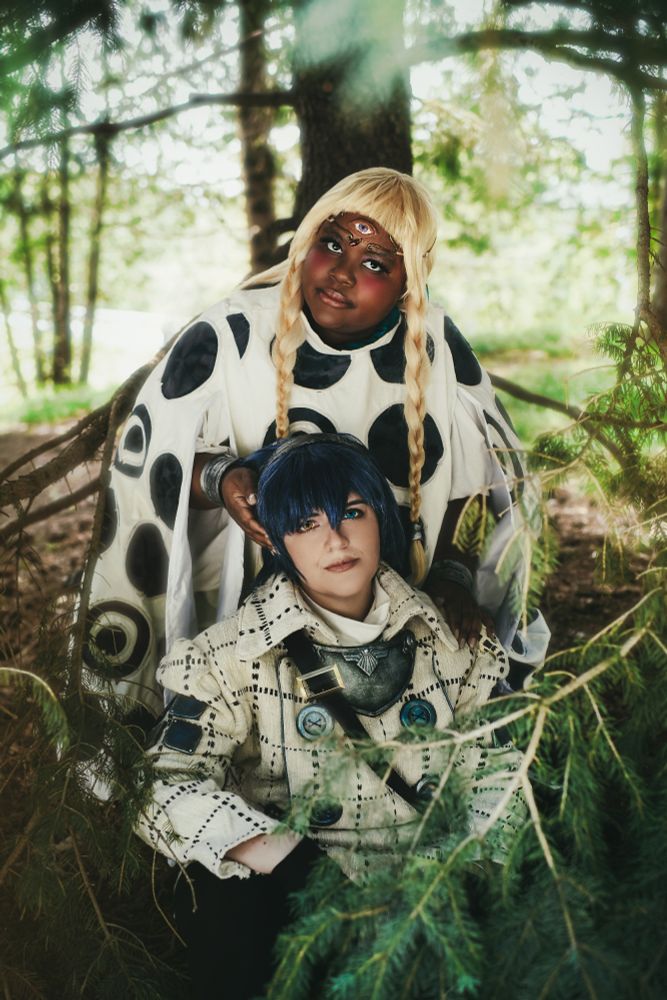two cosplayers dressed as Eupha, a girl in a black and white dress with blonde hair and a third eye, and Will, in a white double breasted coat with blue buttons and a sword, from Metaphor: re fantazio. Eupha is standing behind Will as he sits, cradling his head, surrounded by trees.