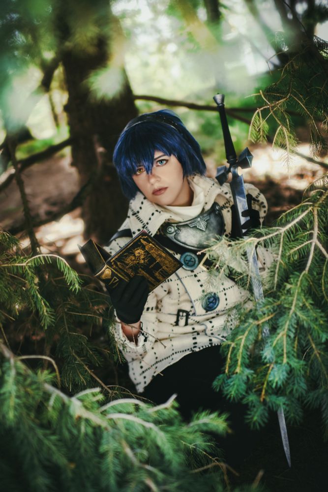 a cosplayer dressed as blue-haired will/the protagonist from metaphor re:fantazio in a white plaid coat with blue accents, leggings, and boots. they're sitting in a pine tree holding a black and gold book and a silver-blue sword, looking at the camera.