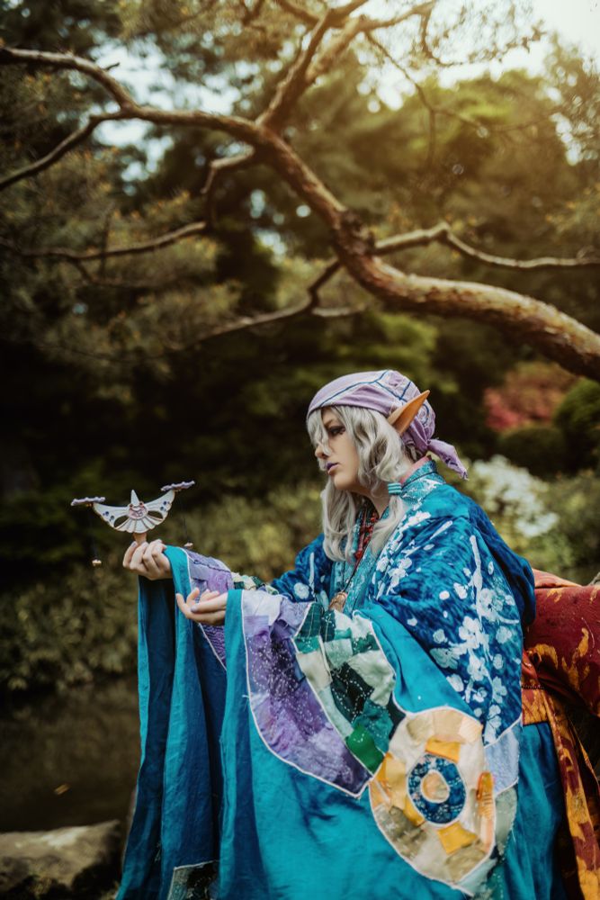 a cosplayer dressed as medicine seller from mononoke in a japanese garden, holding a prop version of the scales. they're dressed in a patchwork and hand painted and dyed kimono