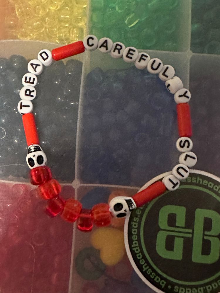 A kandi single. It has various red beads, two skull beads, and the words “tread carefully, slut” on it. 