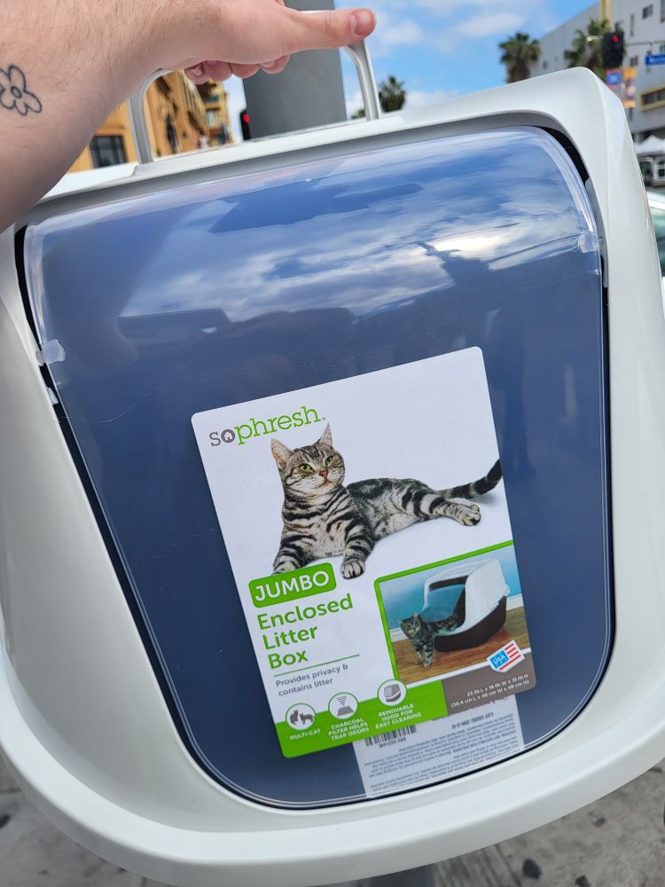 Close-up photo of a jumbo size litter box. I am holding on to the handle, you can only see my hand and bits and pieces of buildings and trees behind the litter box.