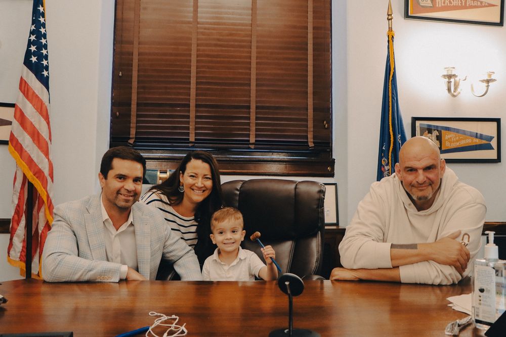 Inside his Washington D.C. office, Senator Fetterman takes a picture to the right of Dominic, a little boy who is smiling and sitting in his chair holding a gavel in the air. Dominic’s parents are also on the left side.