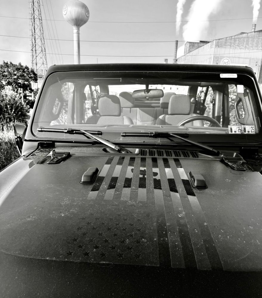 A black and white picture of Senator Fetterman’s union-made jeep with an American flag decal on the hood, in front of the Edgar Thompson Steel Works in his home of Braddock, PA.