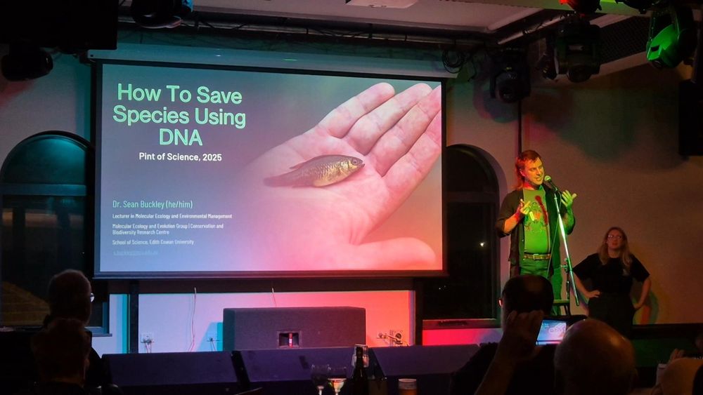 Me, standing on a lit stage next to a large projector screen of the title slide of my presentation, entitled "How to Save Species Using DNA" and featuring a photo of a southern pygmy perch held in a hand. Photo credit: Cass Rowles.