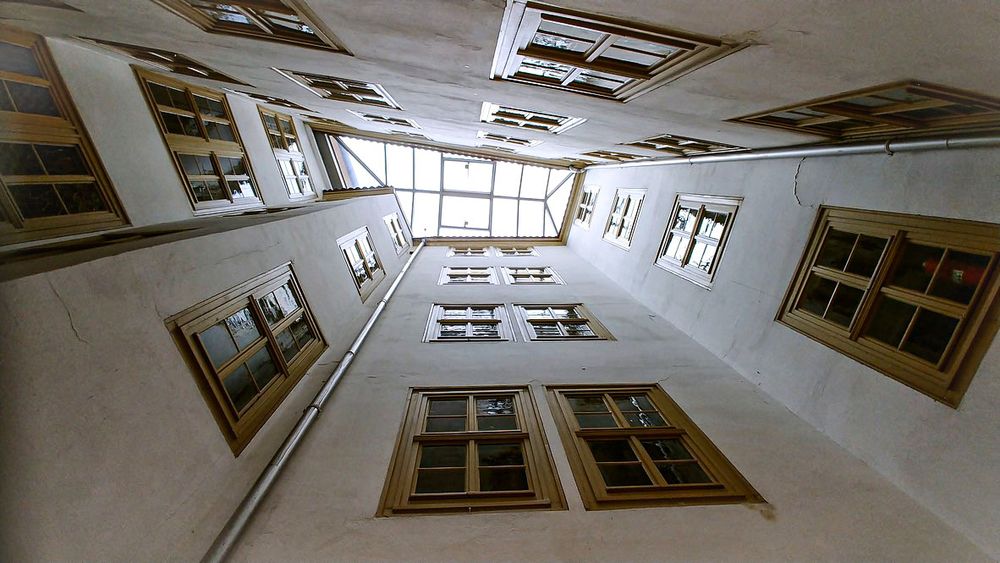 Blick senkrecht nach oben in einen sehr schmalen, hohen Innenhof mit weiß verputzten Wänden. Zahlreiche rechteckige Holzfenster mit Kreuzsprossen befinden sich auf allen vier Seiten in mehreren Etagen übereinander. Ganz oben schließt der Hof mit einem verglasten Dach ab, durch das Tageslicht einfällt, wodurch der Raum hell, aber eng und schachtartig wirkt.