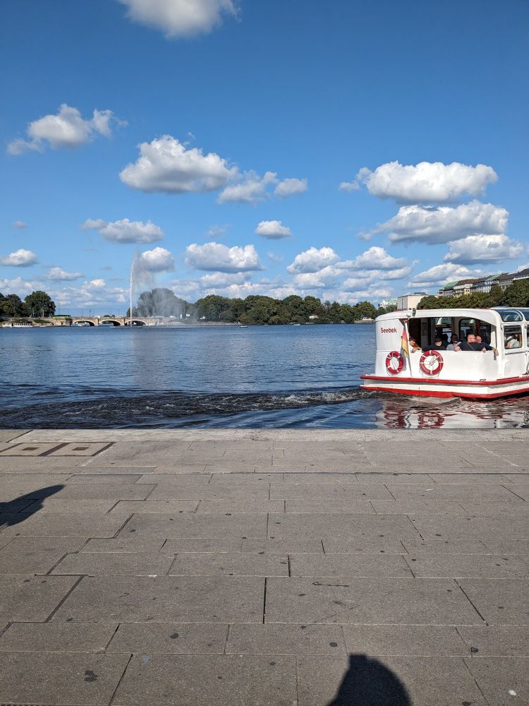 Alster Anleger Jungfernstieg.
Blauer Himmel, ein paar weiße Wolken.
Ein Alsterschiff ist von hinten zu sehen.