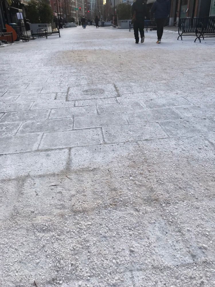 A pedestrian plaza with people walking. The ground is covered with salt, some areas a half-inch thick, like gravel.