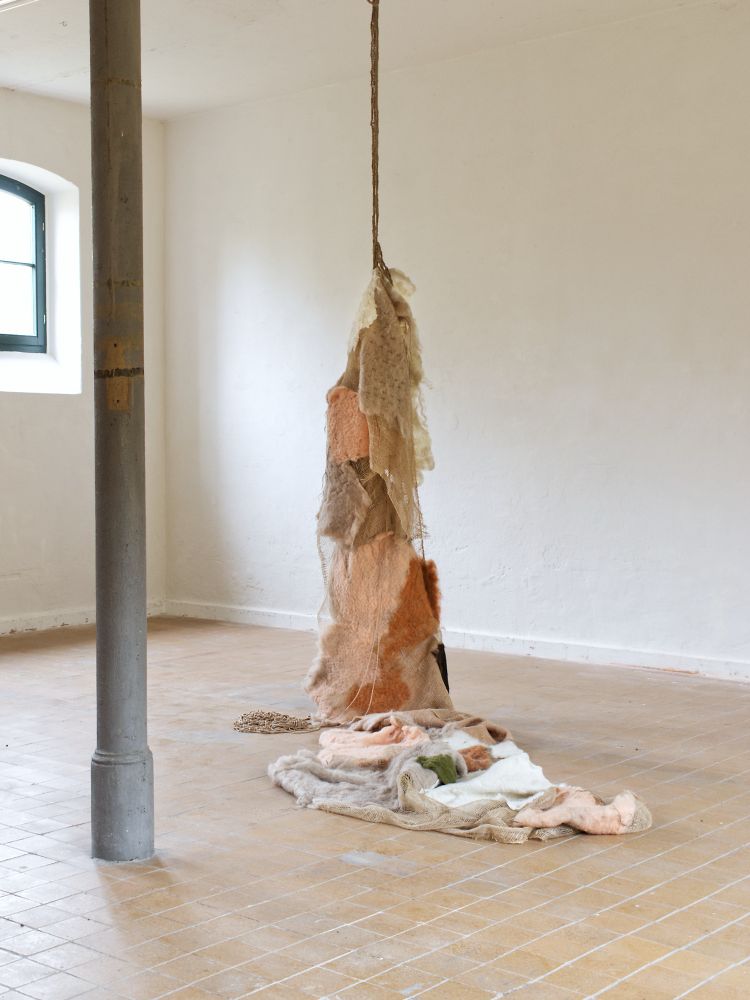 Blick in den Ausstellungsraum des Kunstvereins Röderhof in Sachsen-Anhalt. Man sieht eine Installation aus textilem Material, die von der Decke herabhängt und sich auf dem Boden fortsetzt. Die Farben der Arbeit greifen die der Umgebung auf: hellbraun, rosé, orange und naturweiss. Die Form des Arrangements lässt auch an eine (verhüllte) Figur denken. Man sieht architektonische Elemente: eine stützende Säule, ein Fenster im Anschnitt und gekachelten Boden.