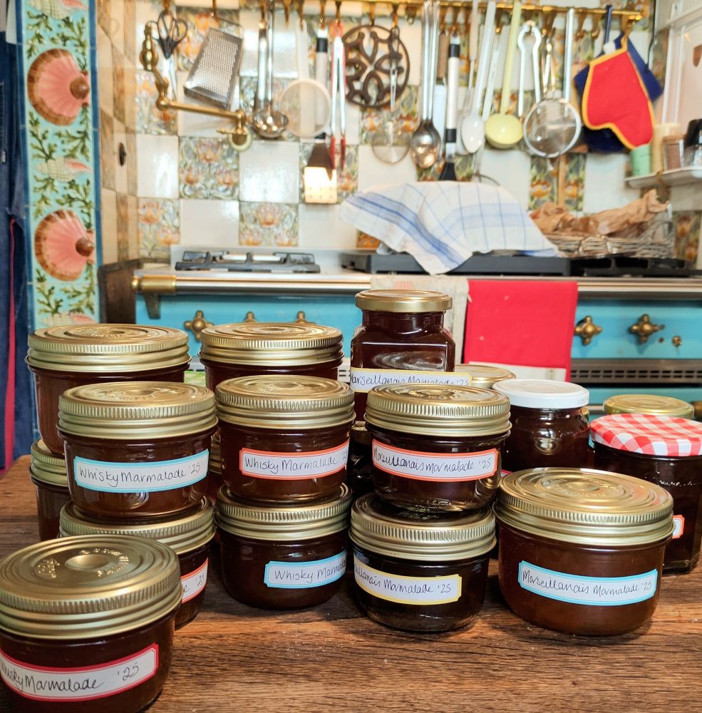 A dozen or so jars of marmalade on a wooden kitchen table, a blue stove and brightly patterned tiles behind. 