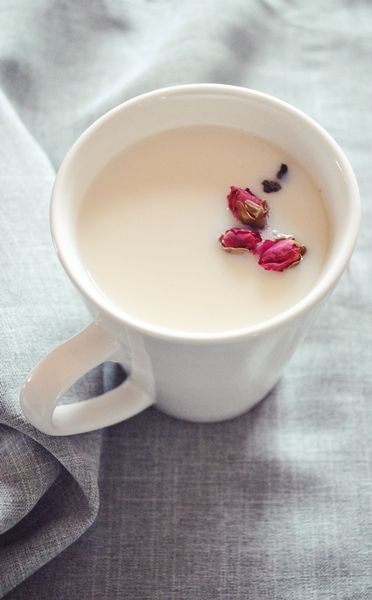 A white ceramic cup full of milky tea or coffee with a few dried rose blossoms floating on top.