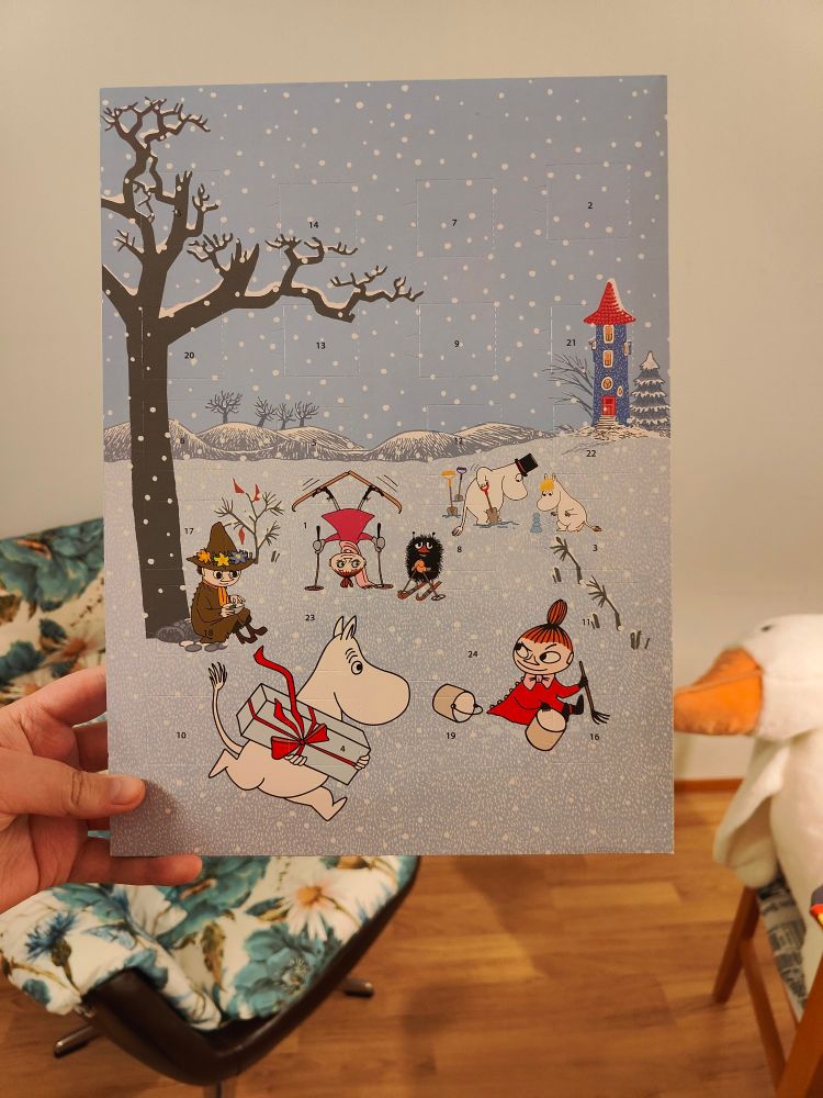 Holding my Moomin-themed advent calendar. The art on it features multiple Moomin characters (sorry I only know them in finnish: Nuuskamuikkunen, Haisuli, Muumipeikko, Muumipappa, Niiskuneiti and Pikkumyy twice for some reason). The characters are outside playing in the snow.
