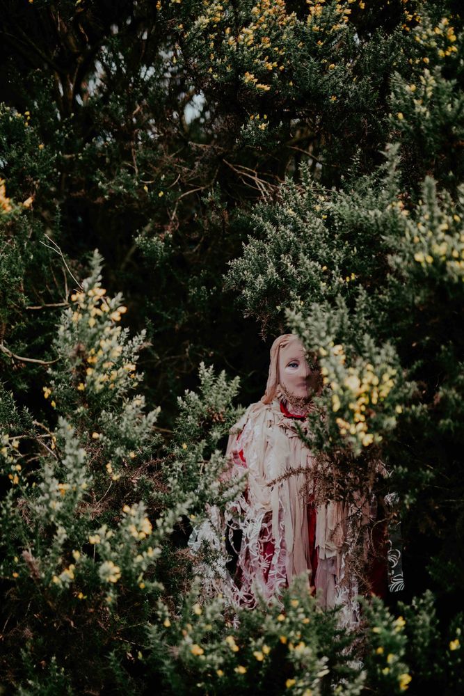 A still from Amanda Coogan’s work The Wren, showing a mysterious figure standing half-hidden amongst trees and flowering brush. The figure is wearing a mask of tights over their head, with holes cut for their eyes, and an elaborate garment with hanging ribbons of many different styles of fabrics.