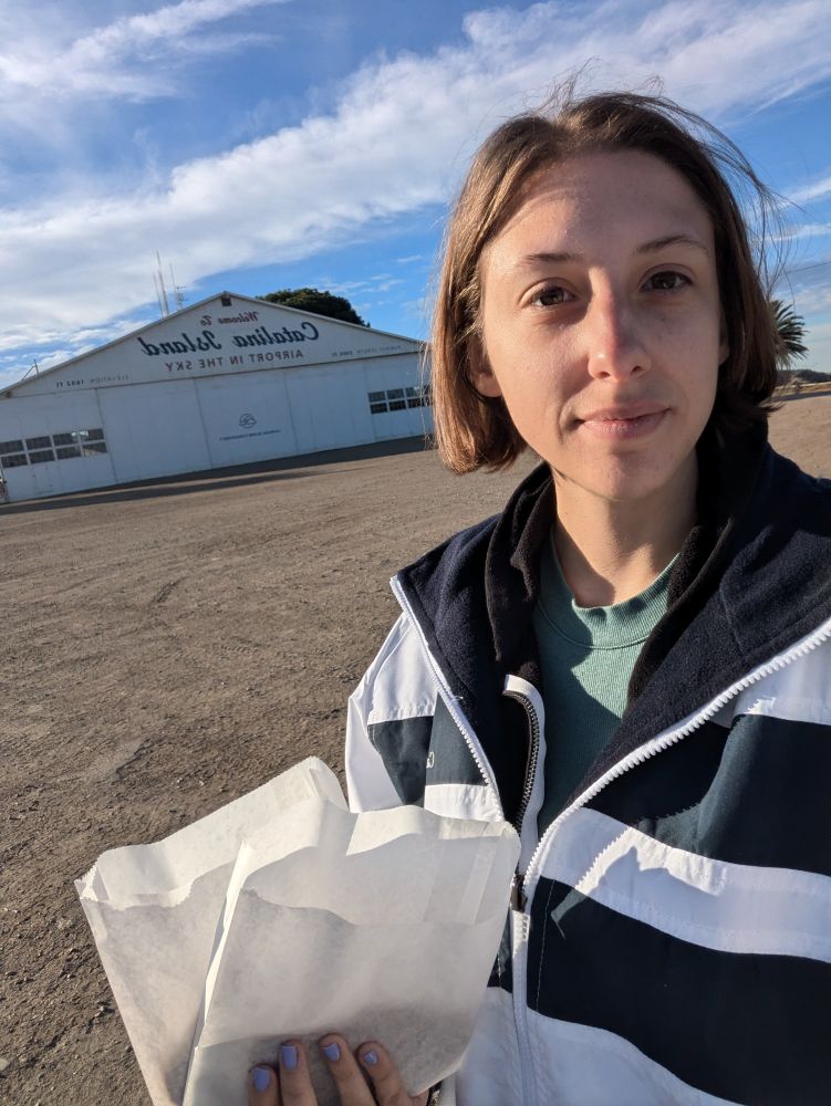 A woman standing outside holding a white paper bag, with a hangar in the background that reads "Welcome to Catalina Island Airport in the Sky."