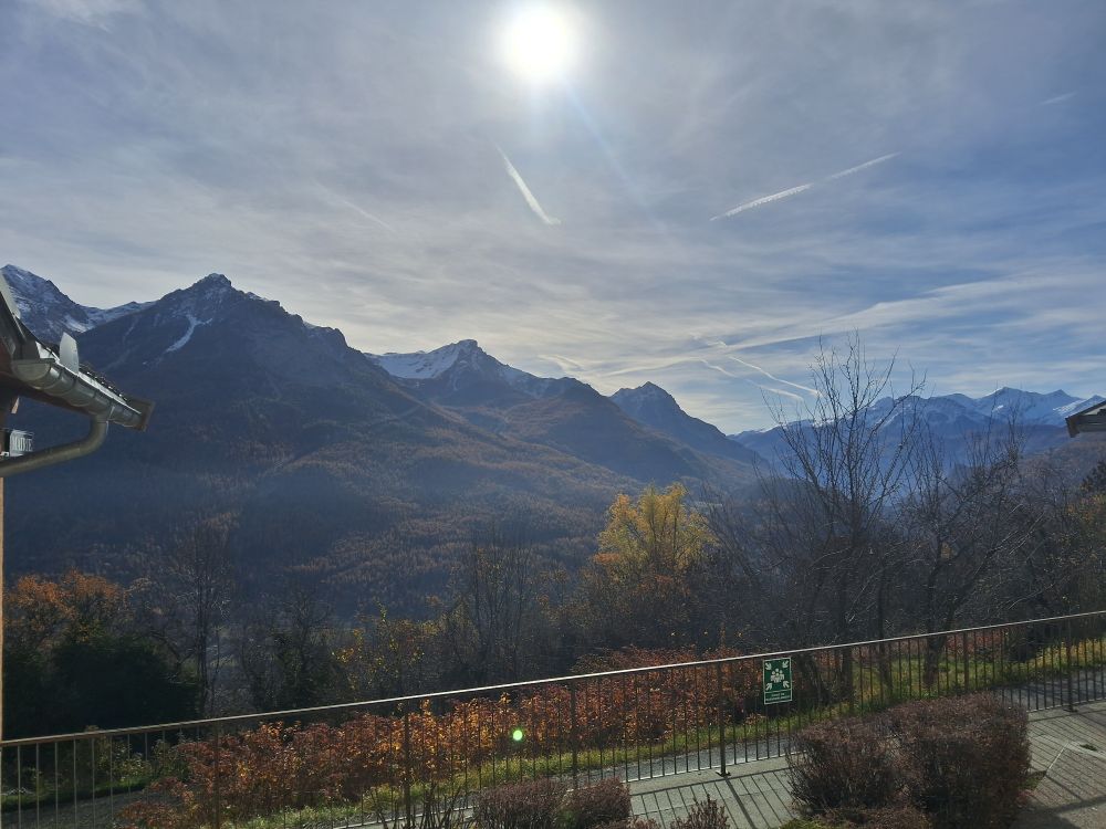 Vu depuis une terrasse : la route juste devant, une vallée, une montagne découpée aux pentes boisées avec de la neige au sommet, un ciel bleu voilé, le soleil 
