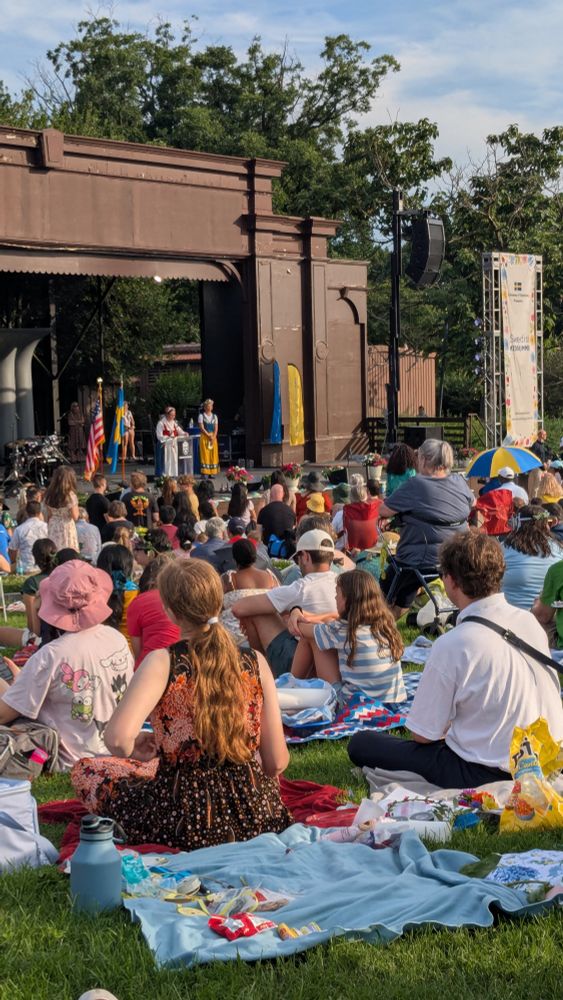 Photo of the Sylvan Theater stage in Washington DC, shot from the lawn facing the stage. A crowd of people sitting on blankets and chairs watches three speakers in traditional Swedish midsummer costume 