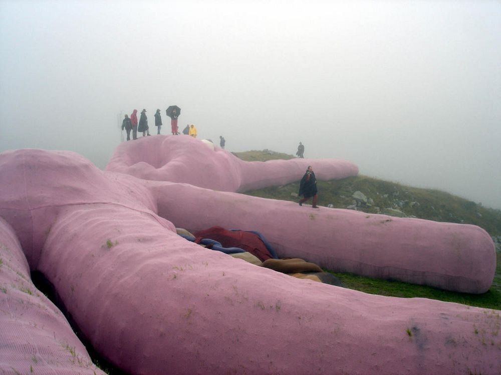 People walk on the surface of a huge knitted rabbit on a foggy mountainside. They are Lilliputian in scale on the giant pink limbs