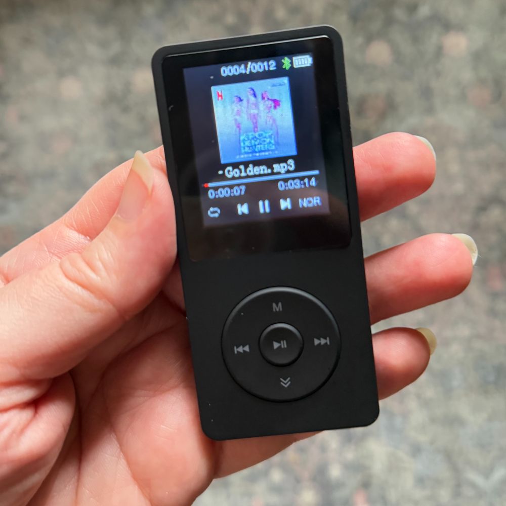 A hand holding a knockoff iPod Nano. It has a small screen showing an mp3 of Golden from Kpop Demon Hunters playing