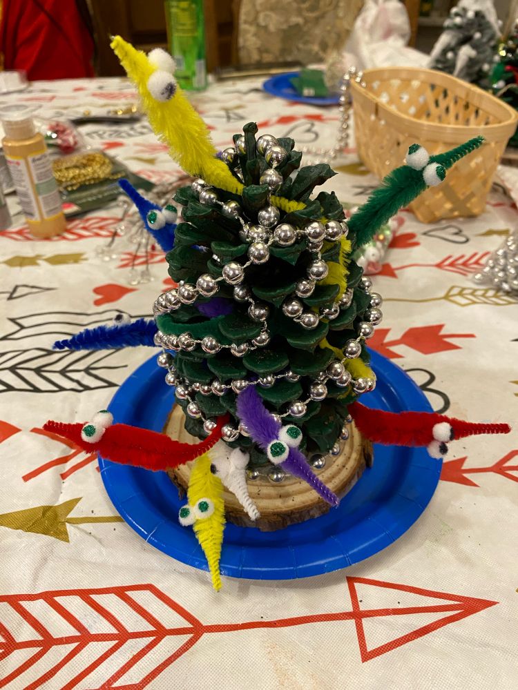 A green painted pinecone on a wooden base. It’s wrapped in silver beads and pipe cleaner that look like the “worm on a string” toy. 