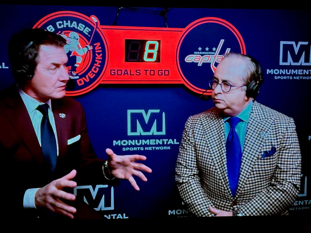 Caps TV announcers Alan May and Joe Beninati sit in the booth at a home game. Behind them, a Gr8 Chase board shows Alex Ovechkin needs to score 8 goals to pass Wayne Gretzky and take sole possession of the career goals record.