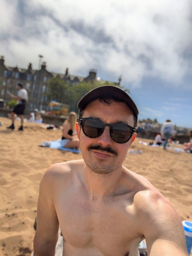 Kevin is at the beach, he is wearing sunglasses and sitting on the sand.