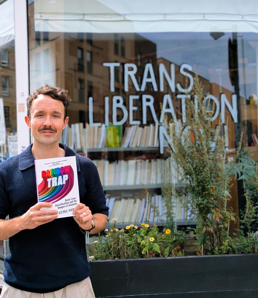 Kevin holding a copy of Rainbow Trap outside the front of Category Is Books. The sign says: Trans Liberation Now!