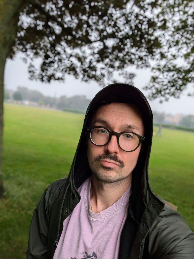 Kevin is in the Meadows in Edinburgh, it is raining and very overcast. He is wearing a rain jacket.