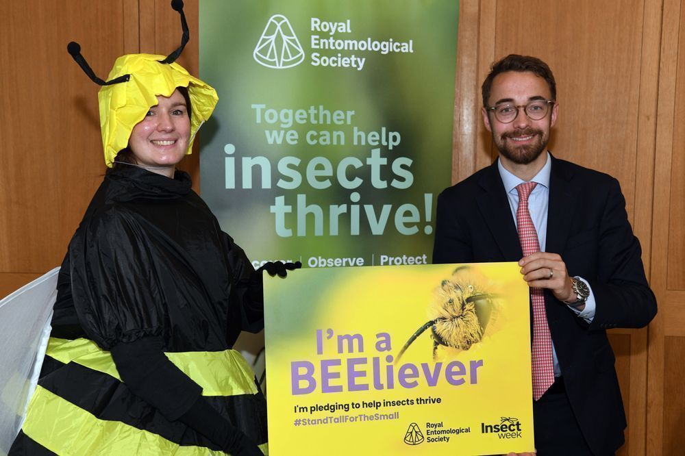 Luke and someone dressed up as a bee with a sign that says "I'm a BEEliever" and a banner in the background