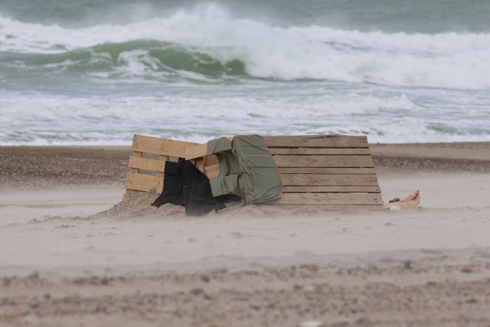 'Skulptur' på stranden. Nogen har stablet paller og andet træ op til et lille læsted. Et par tæpper giver ekstra læ imod vinden. I højre side af fotoet ses et par bare fødder stikke ud. 