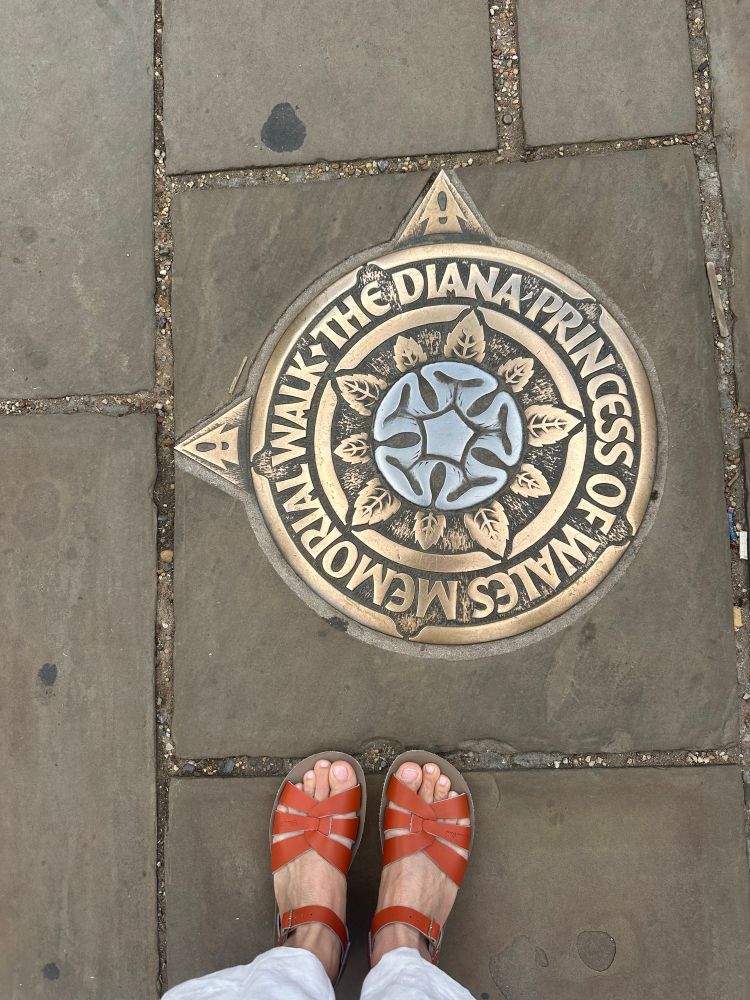 Two feet in orange sandals next to a plaque on the sidewalk that says “the Diana princess of wales memorial walk”