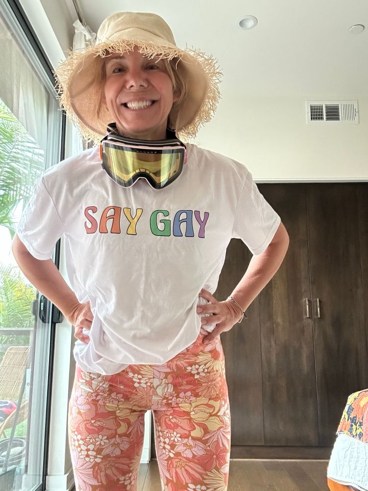 A woman wearing not neutral cooors. She has a straw bucket hat, flower pants, and a SAY GAY T shirt in rainbow colors