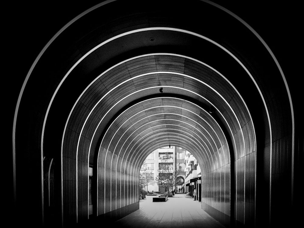 A high contrast black and white photo of a walkway under a building in the shape of a tunnel. A series of Neon tubes go up one side, curve across the ceiling, and back down the other side, receding into the distance. Through the other side, a security guard in a hi-vis jacket can be seen.
