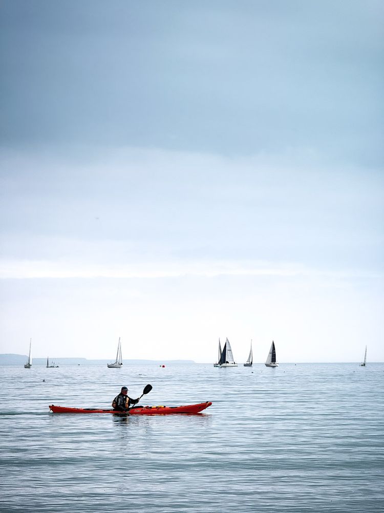 A man in a black and orange wetsuit rowing a red kayak across Weymouth Bay, with about ten sailing dinghies beyond, further out to sea. The kayak has the word 'Leo' painted in black on the hull.