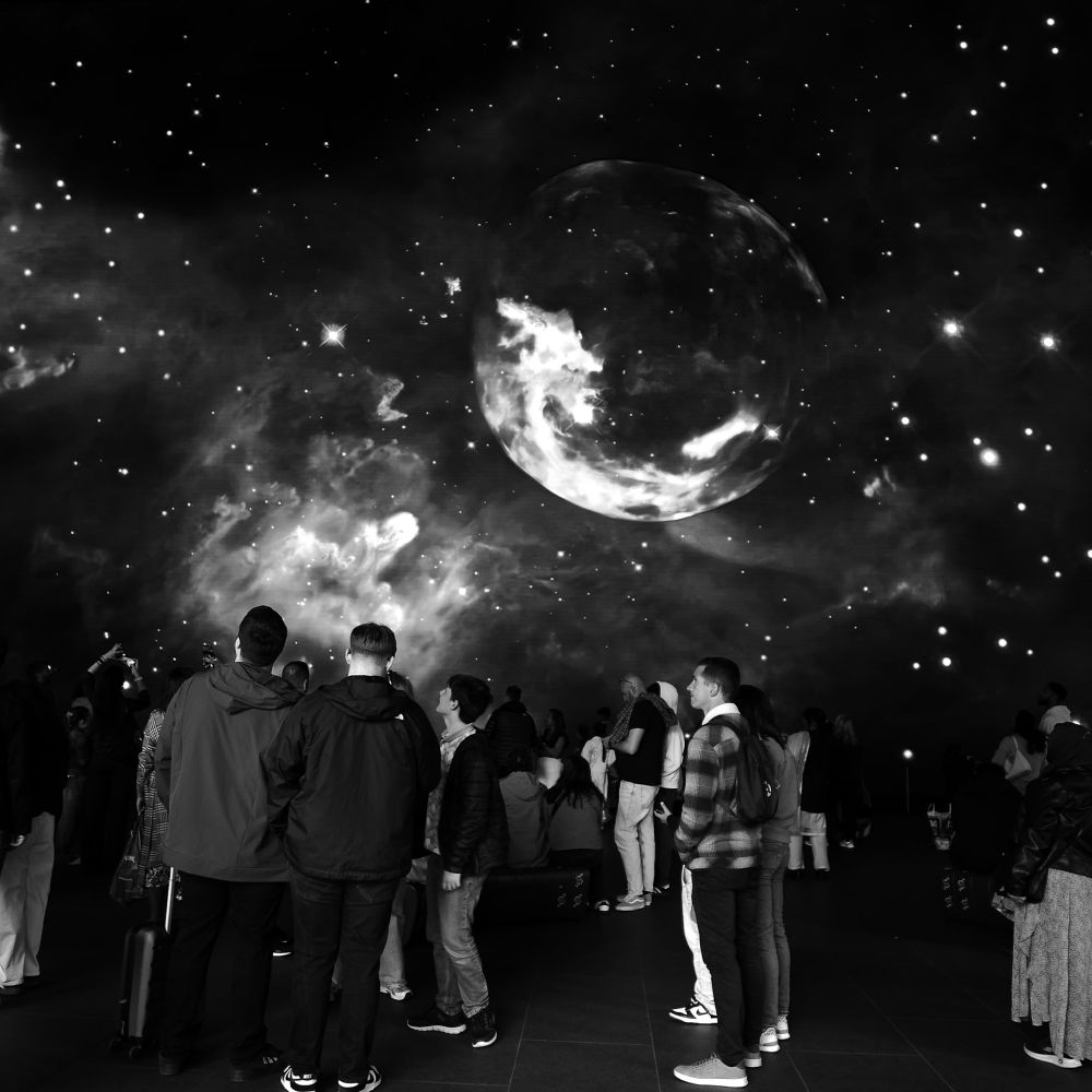People at Outernet in the Now Building in central London, watching an astronomical video on an astronomically-sized video wall. Most of them are facing left or away from the viewer. Above them and slightly to the right is a planet. The rest of the screen is showing stars and cosmic clouds.