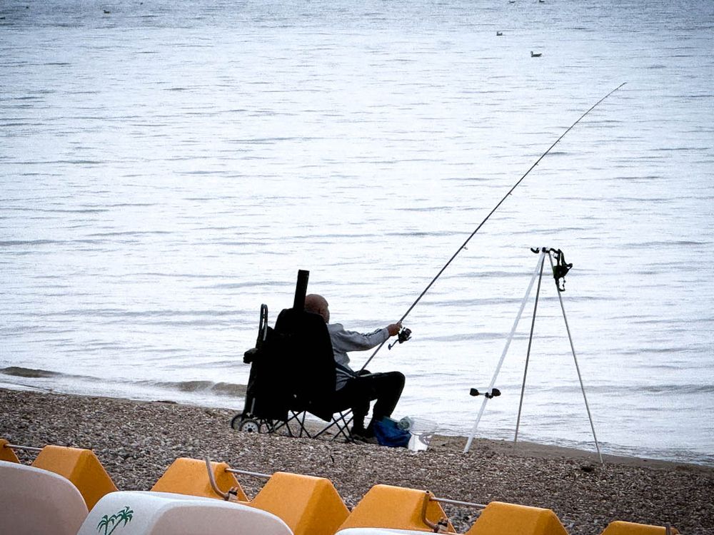 A man sitting on a folding chair, facing out to sea with a wheeled shopping trolley and a fishing rod holder to his left, and a small keep net in a bucket and a tripod to hold the rod to his right. The beach here is very pebbly, and the edges of four yellow and white pedalos can be seen at the water's edge at the bottom of the picture, and a few small seabirds floating near the top.