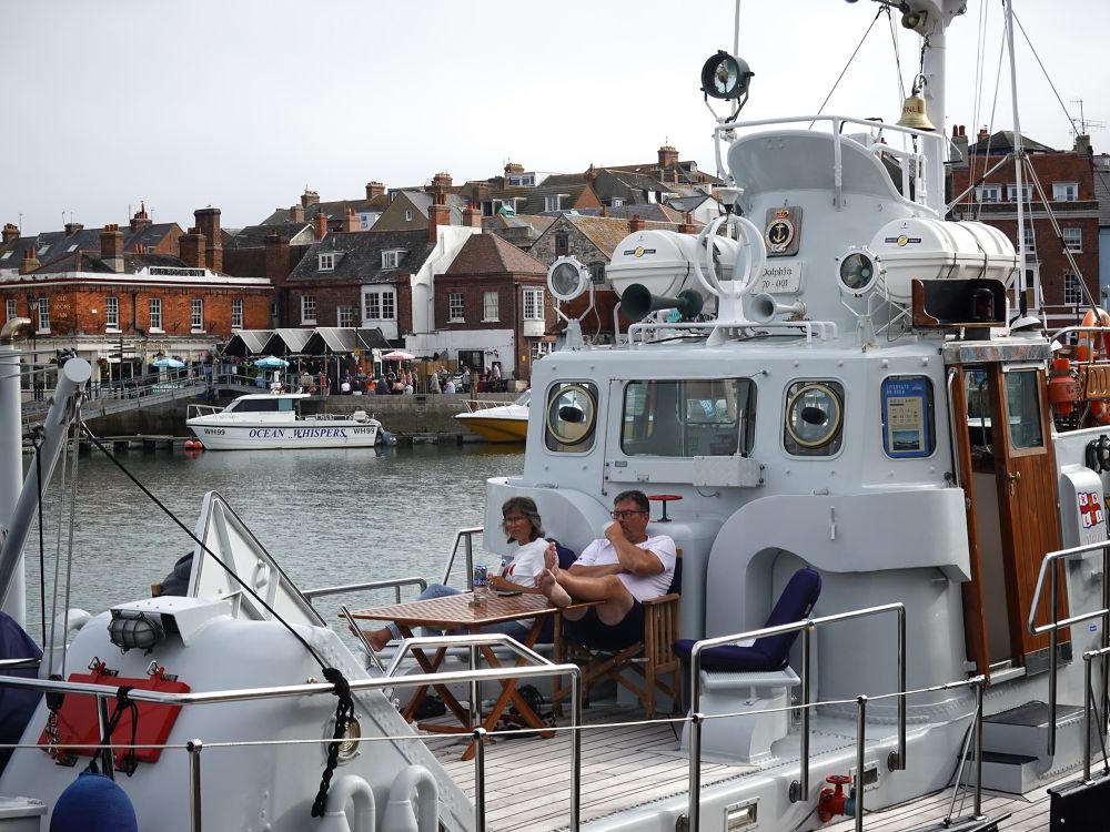 A woman and a man sitting just in front of the wheelhouse of a large white boat in Weymouth Harbour, facing the bow which is beyond the left side of the picture. In front of them is a wooden folding table on which stands a can of lager. Beyond the boat, a part of the harbour can be seen with a much smaller white boat called Ocean Whispers against the opposite side of the harbour. Beyond that is a pub with a couple of dozen people outside, and a number of other buildings.