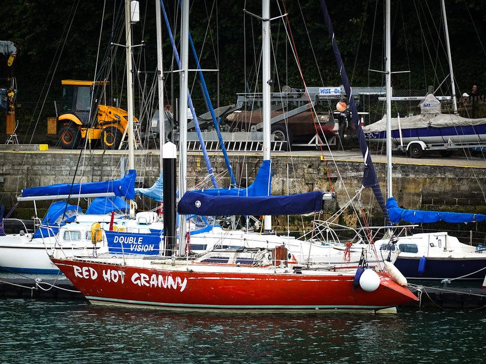 A sailing dinghy in Weymouth Harbour with a red hull and white upper deck, cabin, and trim. The words, "Red hot granny" are painted in large white capital letters along the side of the hull. Two other boats can be seen moored to the same jetty, both white and blue. Another blue and white boat can be seen on a trailer on the harbourside, behind the other boats, along with various pieces of boat-related machinery, including what looks like a small orange tractor.