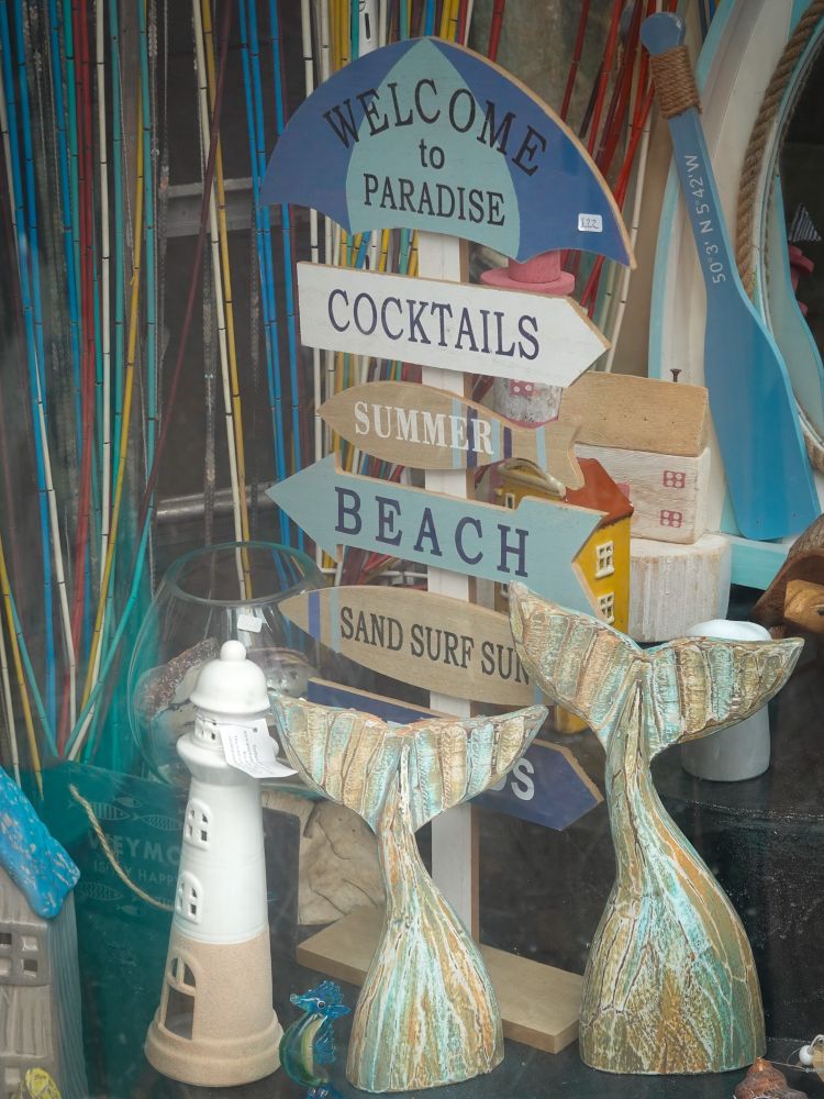 Some of the contents of the window of a seaside gift shop in Weymouth, Dorset, UK, including two wooden 'whale's tails' standing vertically on the shelf behind the window, as if they were sticking out of the sea as the whales dived; a white and brown lighthouse; a couple of small stone cottages; and a tall beach umbrella with the words, "Welcome to Paradise" above a stack of four arrow-shaped signs reading, from top to bottom, "Cocktails", "Summer", "Beach", and "Sand Surf Sun".